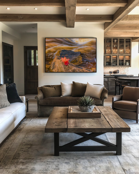 A framed landscape art print featuring a golden sunset over rocky terrain with a red maple leaf, displayed on a white wall in a modern rustic living room setting.