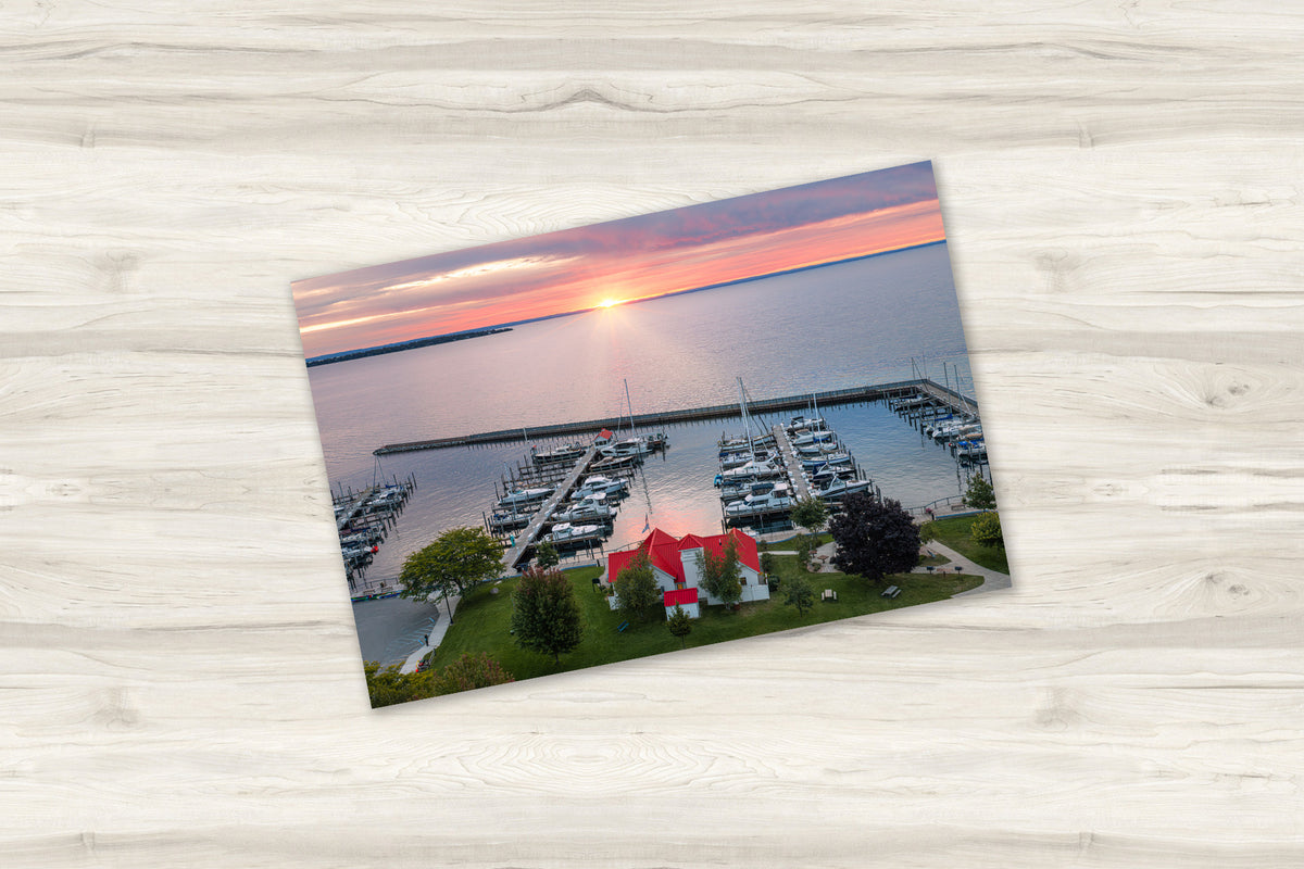 A rectangular landscape photograph print showing a marina at sunset, placed on a light wood-grain surface. The print depicts a harbor with docked boats, green grass, trees, and a red-roofed building under a pink, orange, and blue sunset sky.
