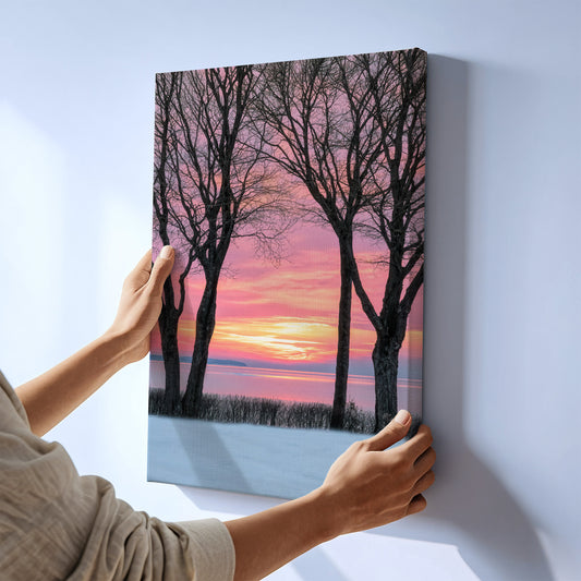 A person holds a vertical canvas wall art print showing bare trees against a pink and orange sunset sky over a snowy landscape, mounted on a light gray wall.