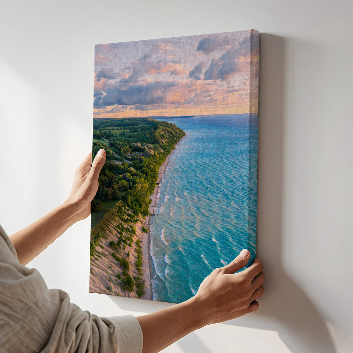 A person holds a vertical canvas wall art print showing an aerial view of a coastal shoreline with turquoise water, green trees, and a cloudy sky at sunset, mounted against a plain light wall.