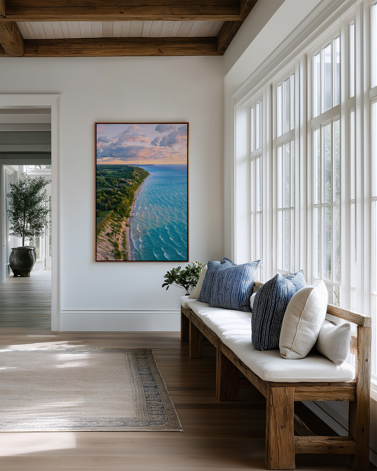 A framed vertical landscape art print depicting Lake Michigan's shoreline with turquoise water and a green peninsula under a cloudy sky, displayed on a white wall in a bright, modern sunroom with a wooden bench and large windows. Aerial view, drone photograph.