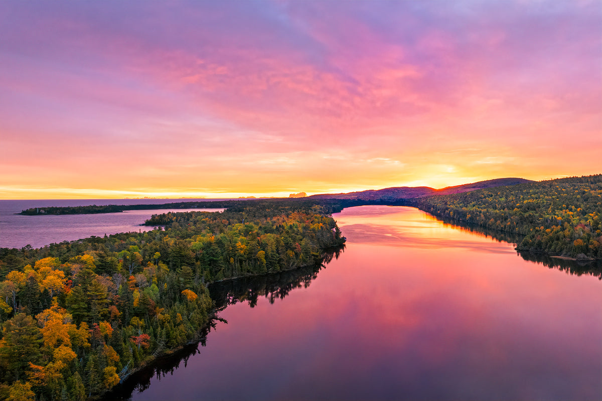 Aerial view of Lake Fanny Houe reflecting a vivid pink, purple, and orange sunset sky, bordered by dense forest with autumn foliage.