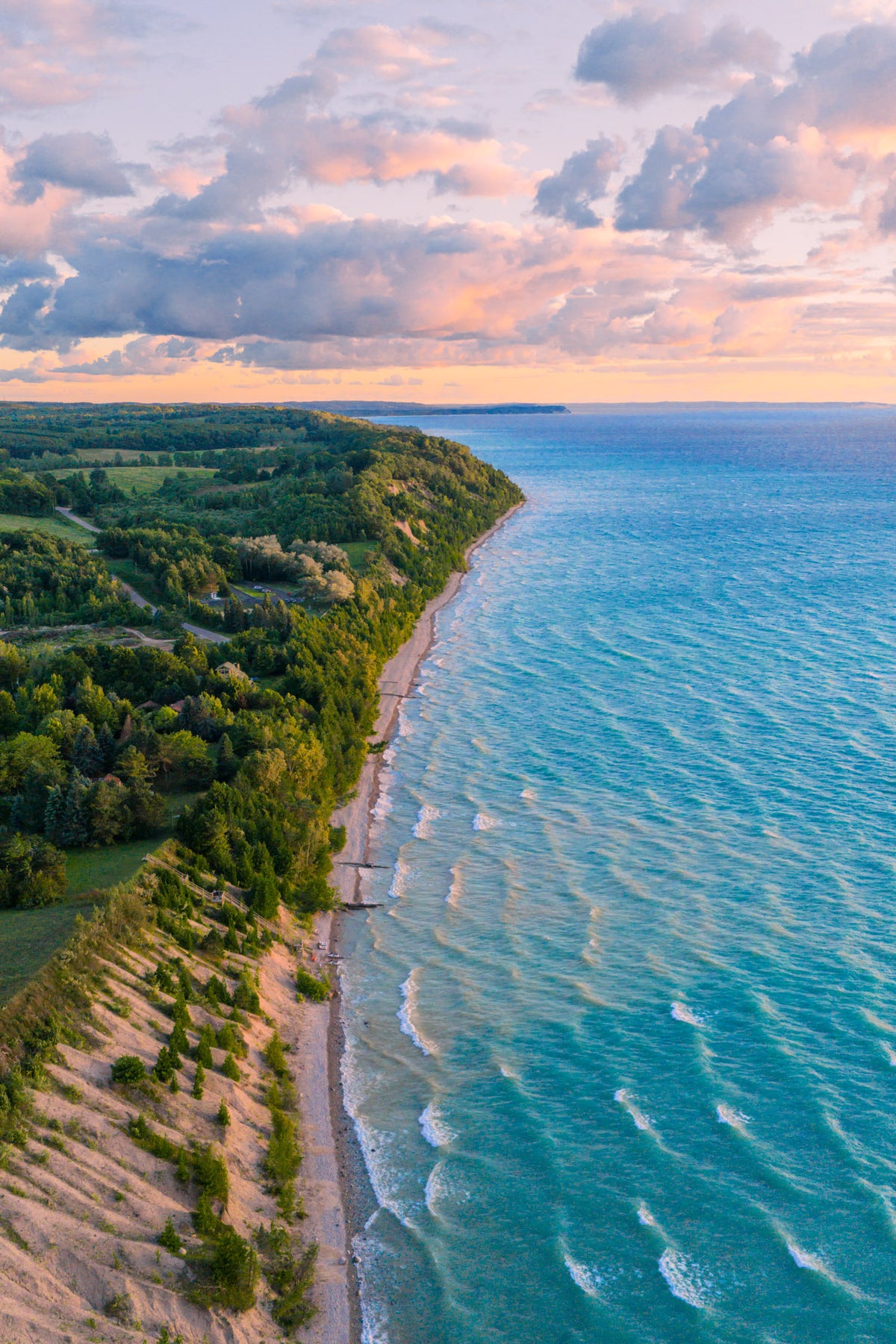 Aerial coastal landscape of Lake Michigan with turquoise blue water, a sandy shoreline, dense green forest, and a dramatic cloudy sky at sunset.