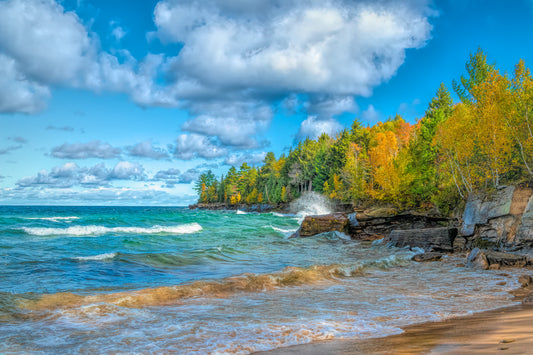 A scenic view of Lake Superior with fall foliage in the background, showing a clear blue sky, white clouds, and a rocky shoreline.