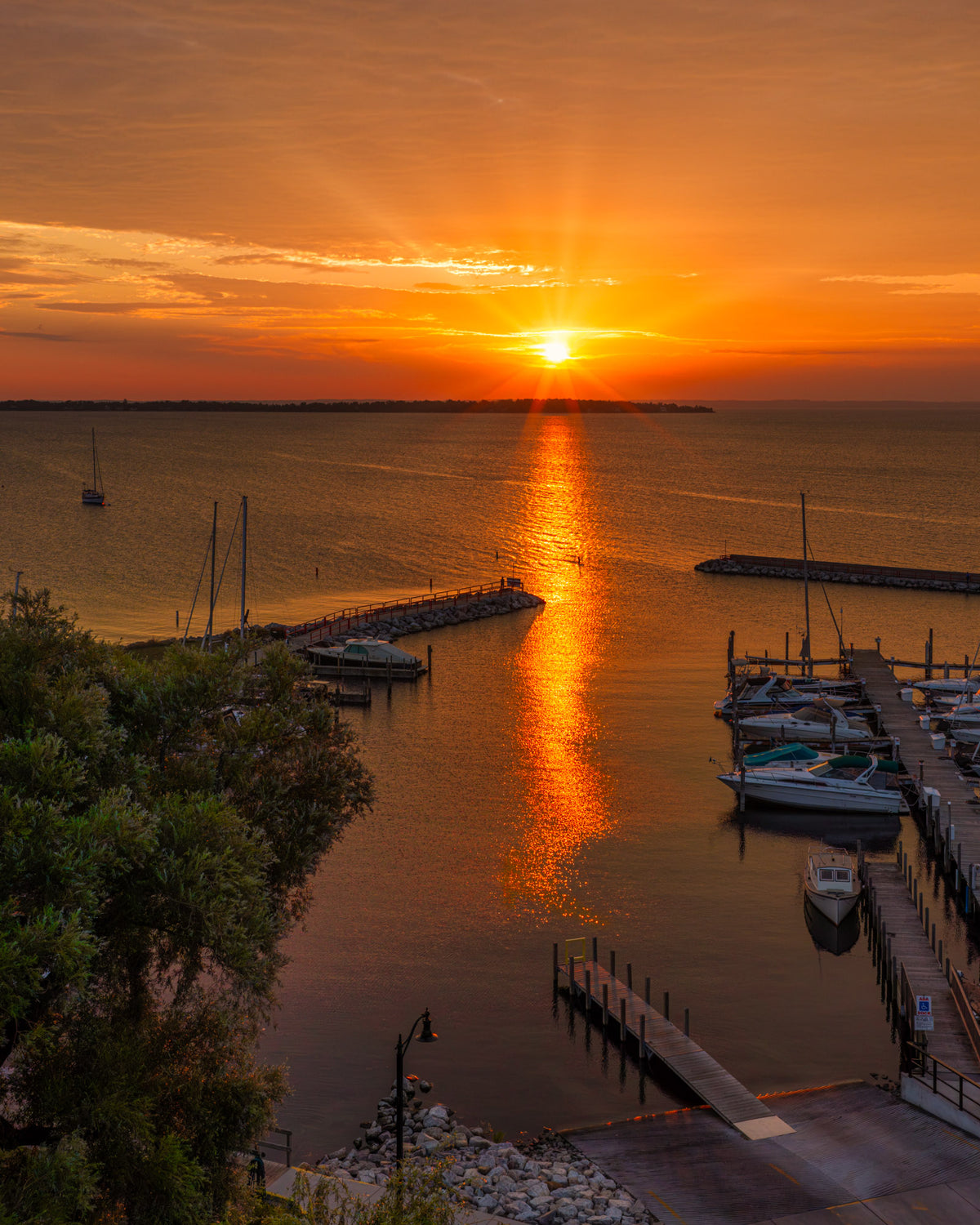 A scenic sunrise over Northport's calm harbor with boats docked at piers, bright sun rays reflecting across the water, and warm orange and yellow tones filling the sky.