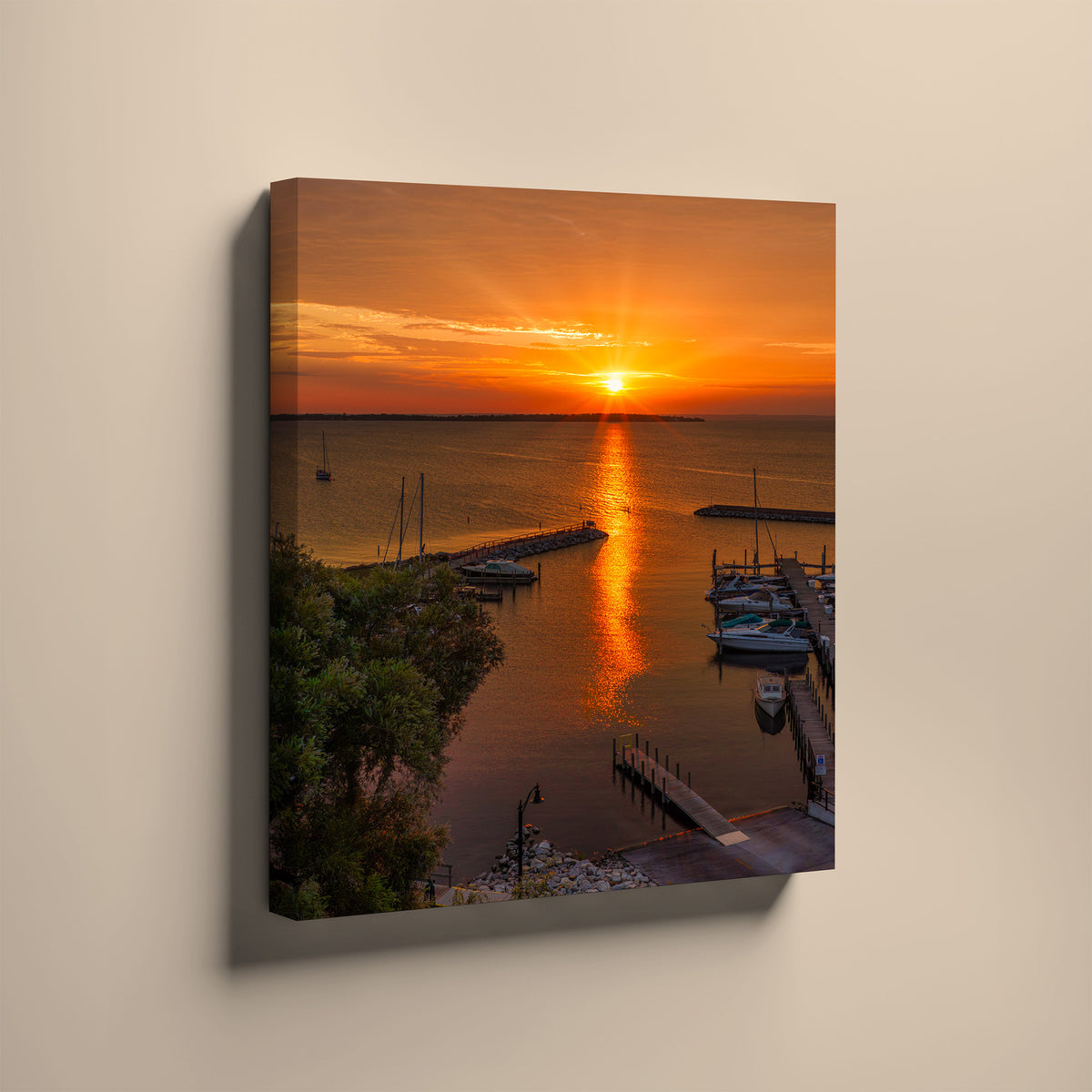 A vertical canvas wall art print depicting a vibrant orange and yellow sunset over a harbor with boats and docks, shown hanging on a light beige wall. Northport, Michigan.  Leelanau County.