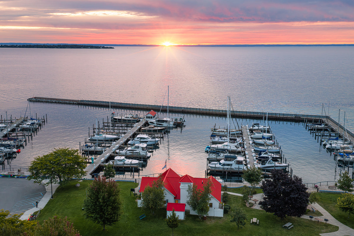 Aerial view of a marina at sunrise over calm water of Grand Graverse Bay, with docked boats, a pier, green lawn, trees, and a small white building with a red roof in the foreground.