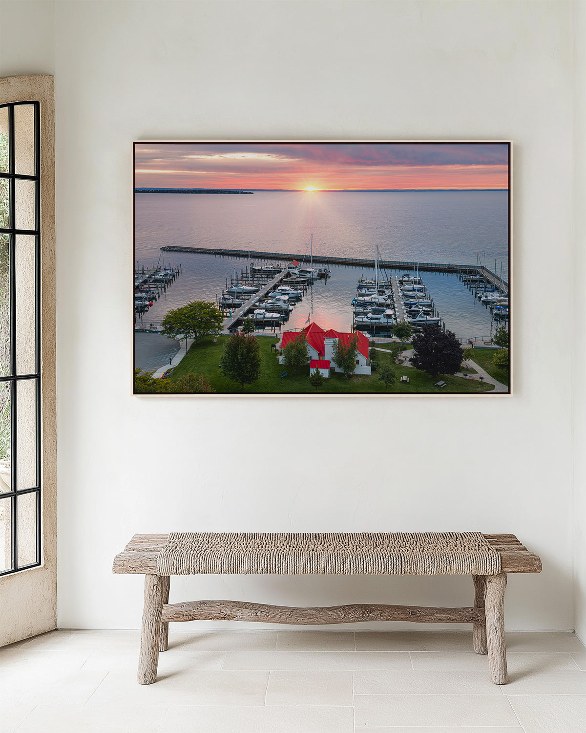 A framed landscape art print of Northport, Michigan marina at sunrise hangs on a white wall above a rustic wooden bench. The print depicts a harbor with docked boats, green grass, and a small red-roofed building under a pink and orange sky.