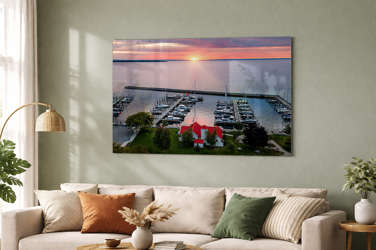 A large metal rectangular wall art print showing a marina at sunset hangs above a beige sofa in a styled living room. The print depicts boats docked at piers, green landscaping, and a pink-orange sky over water. Grand Traverse Bay, Lake Michigan in Northport.