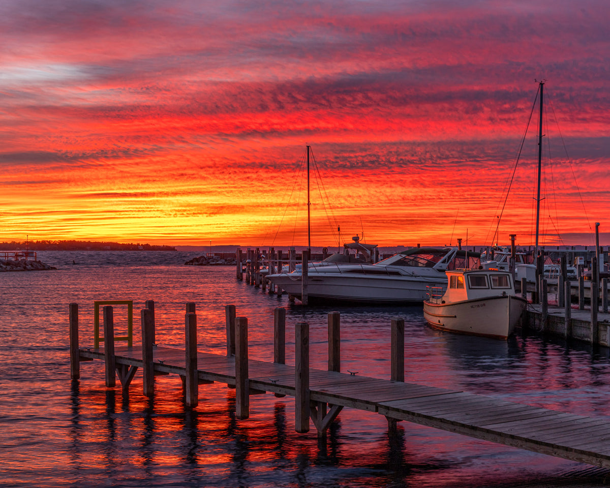 A vibrant sunrise over a marina in Northern Michigan, featuring a bright orange and red sky reflected in the calm water, with wooden docks and several boats docked at the pier.