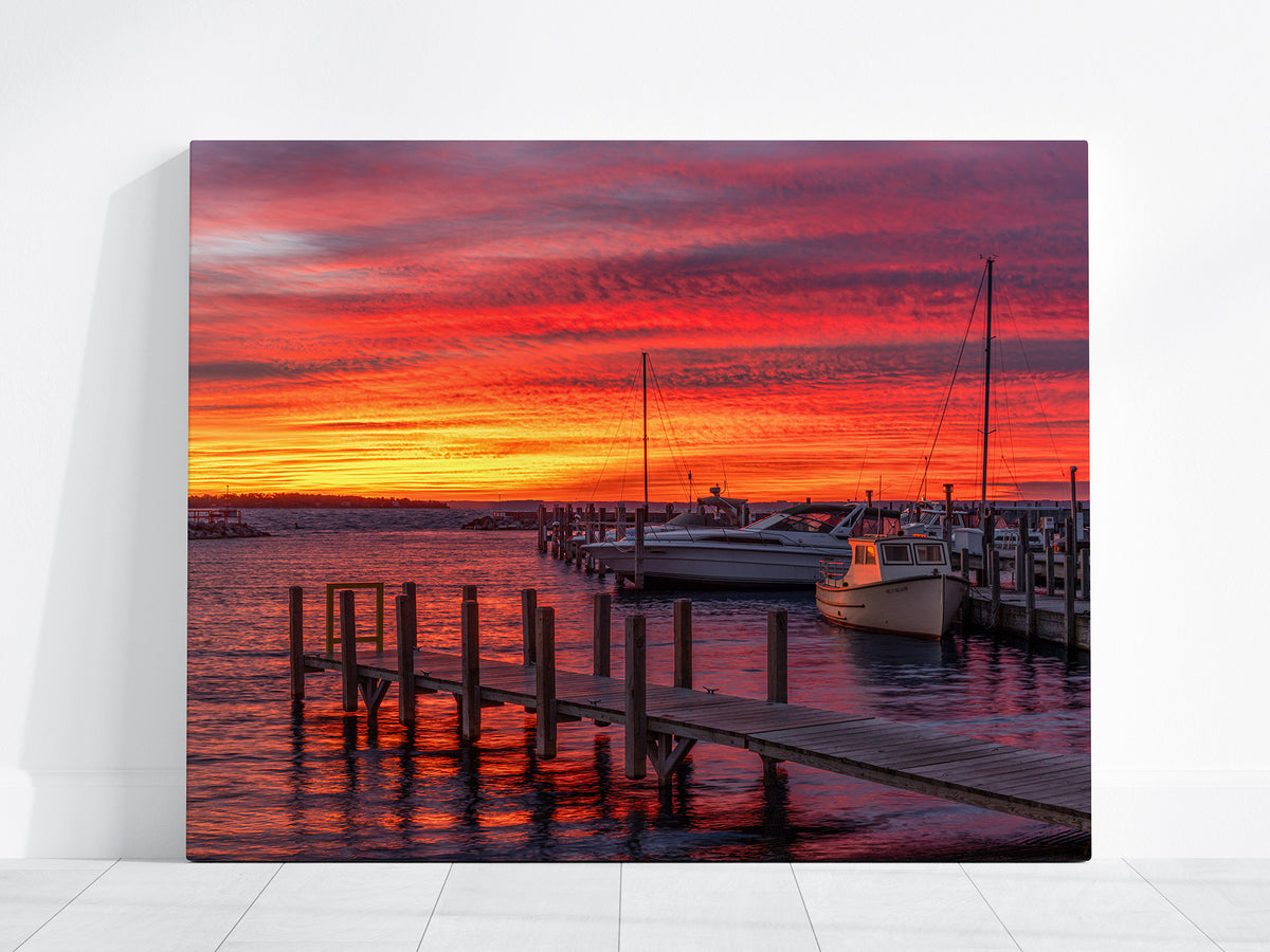 A rectangular wall art print or canvas leaning against a white wall on a light floor, showing a vivid sunrise marina scene with red, orange, and pink sky, boats, and wooden docks reflected in the water. Grand Traverse Bay.