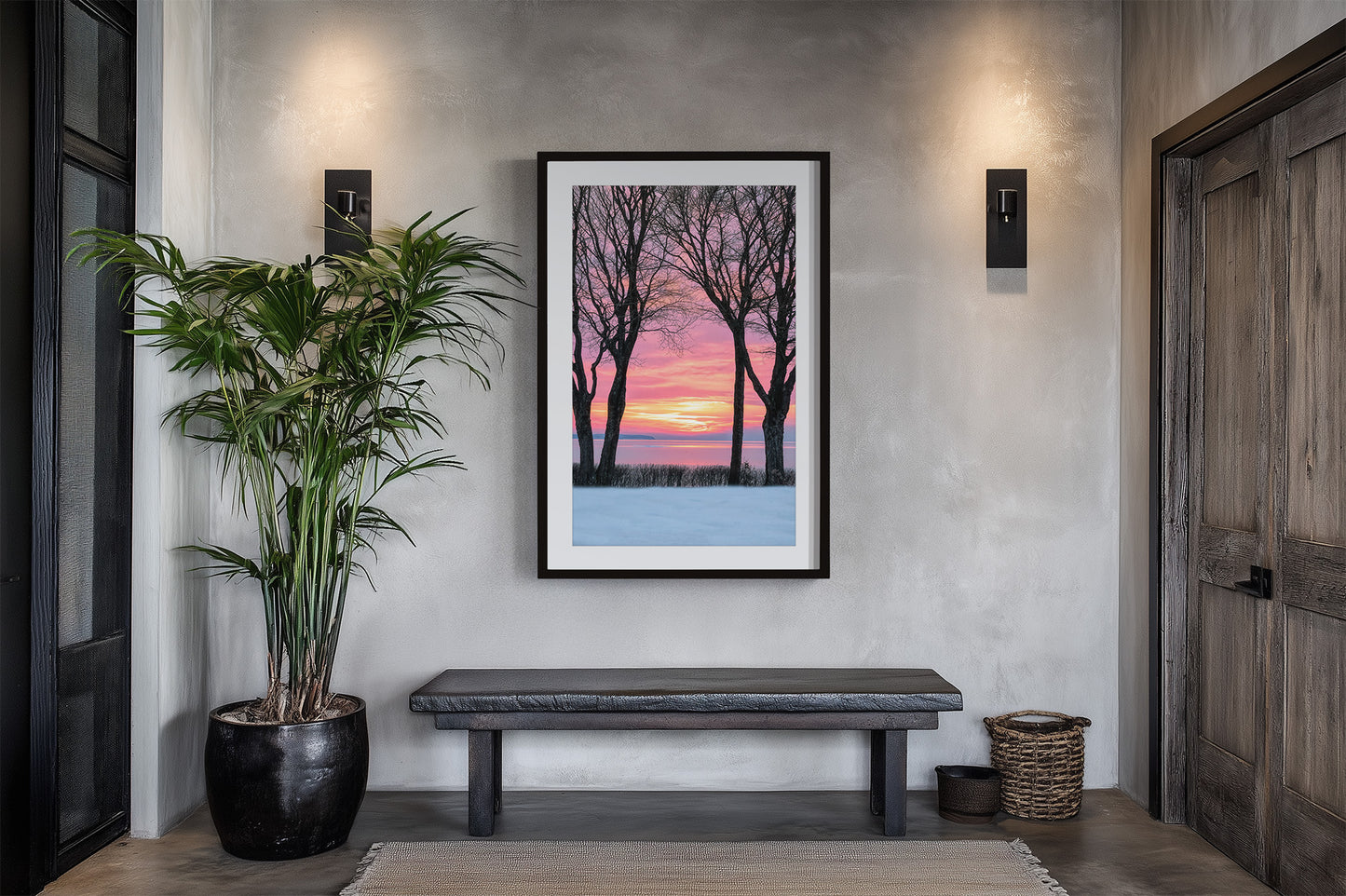 A framed vertical art print depicting a winter sunset with bare trees against a pink and orange sky, displayed on a textured gray wall above a dark bench in a modern entryway.