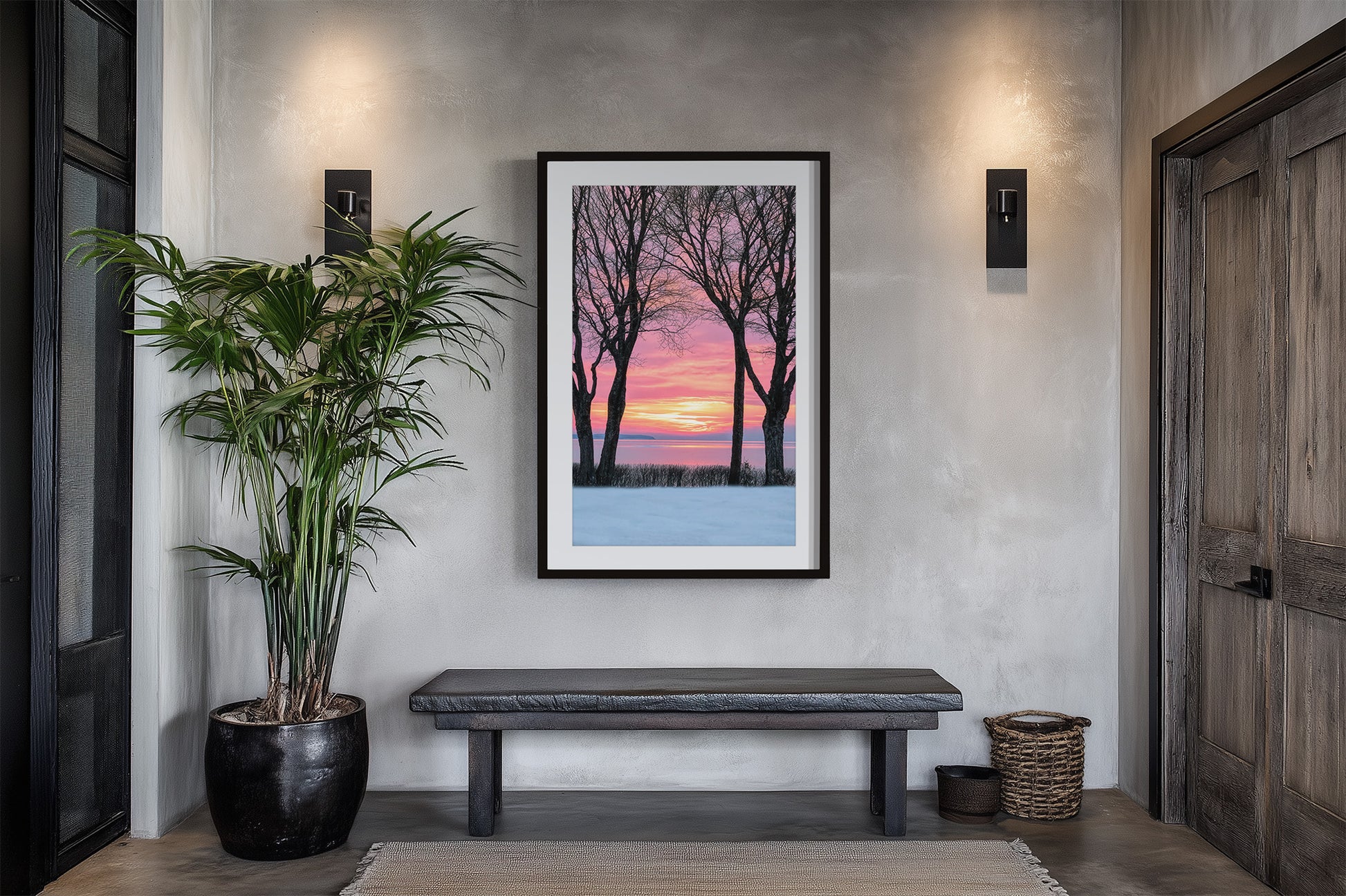 A framed vertical art print depicting a winter sunset with bare trees against a pink and orange sky, displayed on a textured gray wall above a dark bench in a modern entryway.