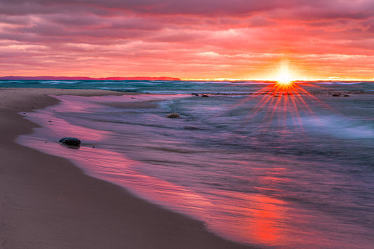 Sunset over a beach with vibrant red, orange and blue colors with glowing light on water. Sunburst is at the horizon.