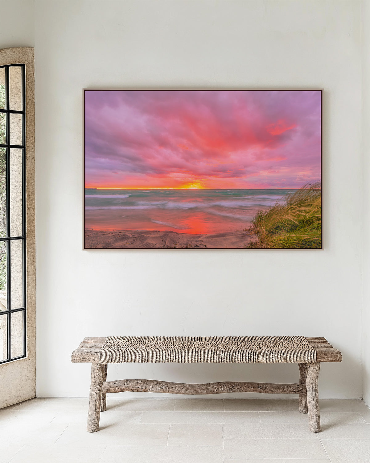 Framed scenic landscape of  Lake Michigan, Van's beach at sunset, featuring dramatic pink and orange clouds, turquoise water, wet sand reflecting the sky, and grass in the lower right foreground over a wicker bench in entry way.
