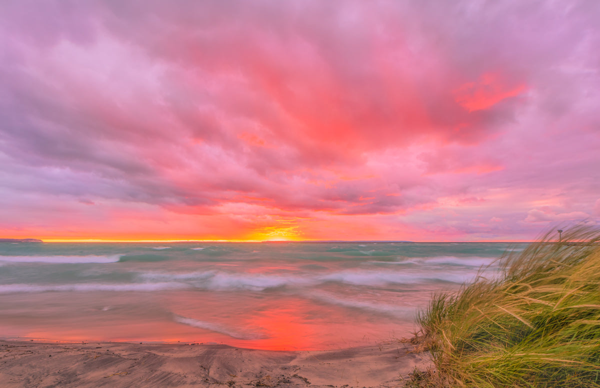 A scenic landscape photograph of a Lake Michigan beach at sunset, featuring dramatic pink and orange clouds, turquoise water, wet sand reflecting the sky, and grass in the lower right foreground.